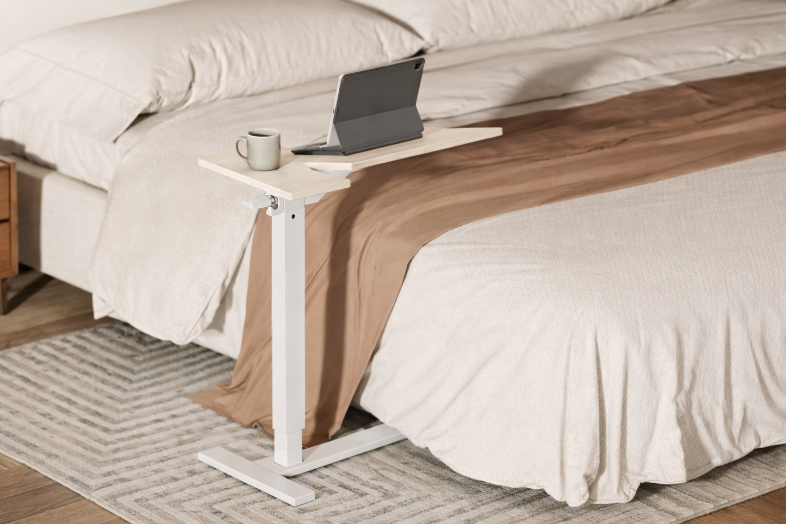 White adjustable C-shaped side table with a light wood desktop, featuring a laptop and mug, positioned next to a bed with neutral bedding.